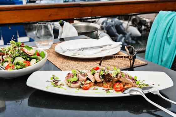 Dekkede restaurantbord med kjøttrett, og frisk salat, med utsikt mot båter på vannet.