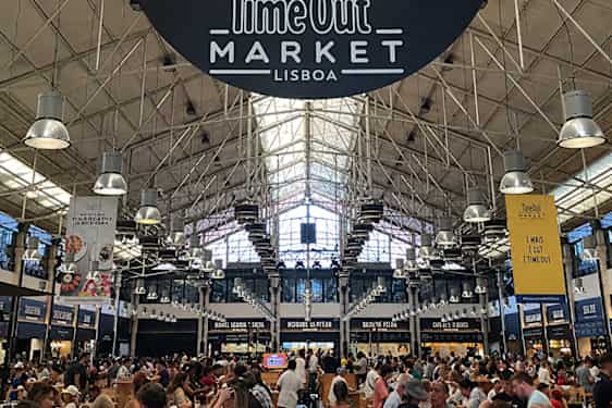 Skiltet «Time Out Market Lisboa» henger over en travel mathall med lange bord, matboder og mange besøkende. Ståldragere, takvinduer og industrilamper preger rommet.