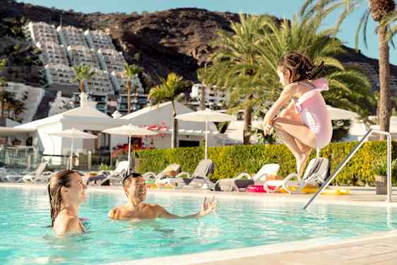 En liten jente hopper i bassenget, hvor foreldrene nyter ferien og slapper av på Gran Canaria