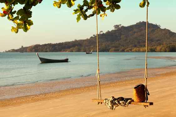 Paradisstrand i Thailand med en huske hengende fra et tre, en båt på vannet og fjell som reiser seg i bakgrunnen.