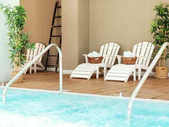 Innendørs svømmebasseng med klart blått vann og stiger i rustfritt stål. Solsenger med håndklær står langs bassengkanten, flankert av grønne planter og lyse vegger som skaper en rolig spa‑atmosfære.