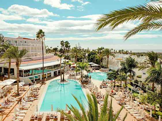 Oversiktsbilde av et resort med stort utendørs basseng i midten, omgitt av solsenger og palmer. Lave hotellbygninger rundt bassengområdet, med sjø og strand synlig i bakgrunnen under blå himmel.