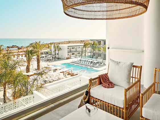 Utsikt fra hotellrom med balkong, stol og bord. Nedenfor ligger et innbydende bassengområde omgitt av palmer og moderne hotellbygninger i solfylt feriemiljø.