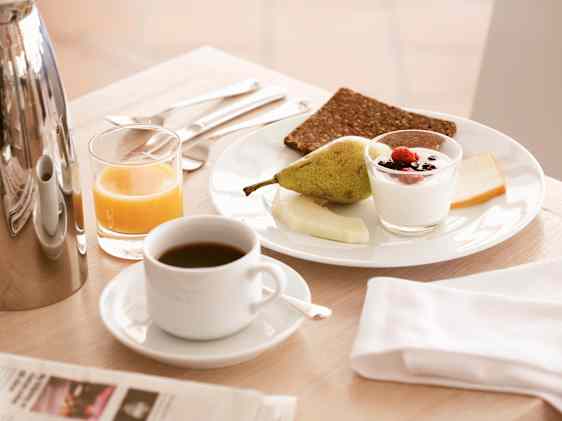 Frokostbord med kaffekopp, glass appelsinjuice, tallerken med pære, yoghurt med bær og brødskiver. Serviett og bestikk ved siden av, med kaffekanne i bakgrunnen på et lyst bord.