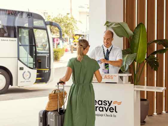 En gjest med koffert sjekker inn bagasje ved hotellets resepsjon, mens en buss står parkert utenfor.