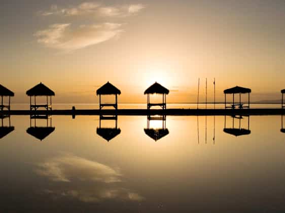 Solnedgang over rolig vann med silhuetter av strandpaviljonger som speiler seg i vannoverflaten.