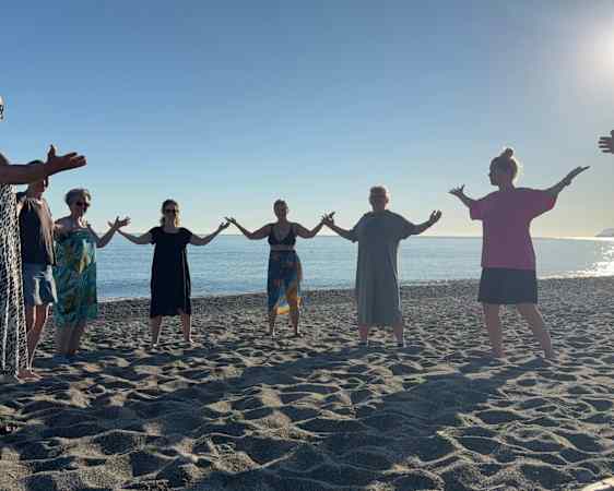 En gruppe mennesker står på en strand med armene utstrakt mot hverandre i solnedgangen.