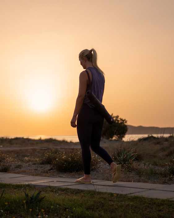 Person går langs en sti i kystlandskap ved solnedgang, med yogamatte over skulderen. Solen står lavt over horisonten og lyser opp havet, vegetasjon og åser i bakgrunnen i varmt, gyllent lys.