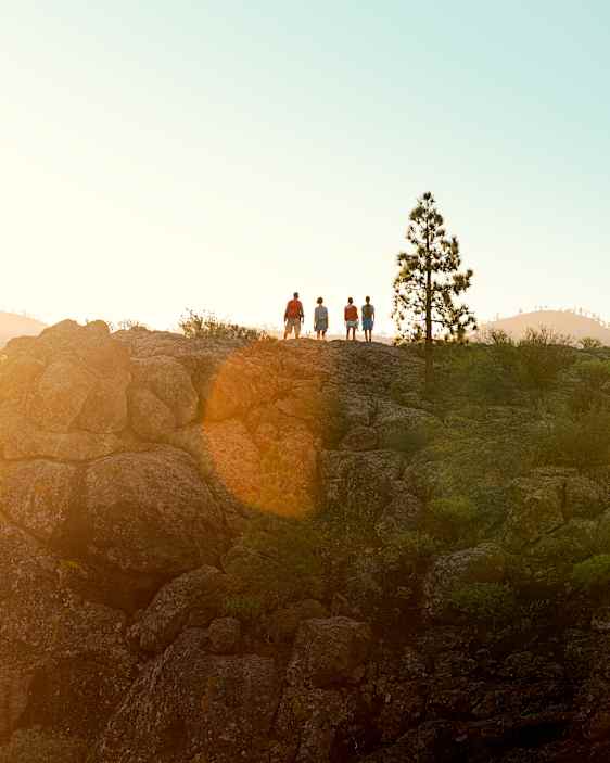 Gruppe personer står på en fjellknaus og ser utover et fjellandskap i varmt sollys. Solen er lav på himmelen og skaper et gyllent skjær over åser, steinformasjoner og trær i bakgrunnen.