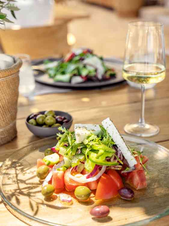 En salat med tomater, oliven, løk, grønn paprika og hvit ost ligger på en glassplate. Et glass og flere retter står i bakgrunnen.