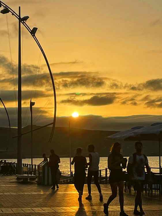 Solen står lavt over havet og farger himmelen gul og oransje. Flere mennesker går langs en promenade med høye, buede lyktestolper, mens mørke skyer ligger over horisonten.