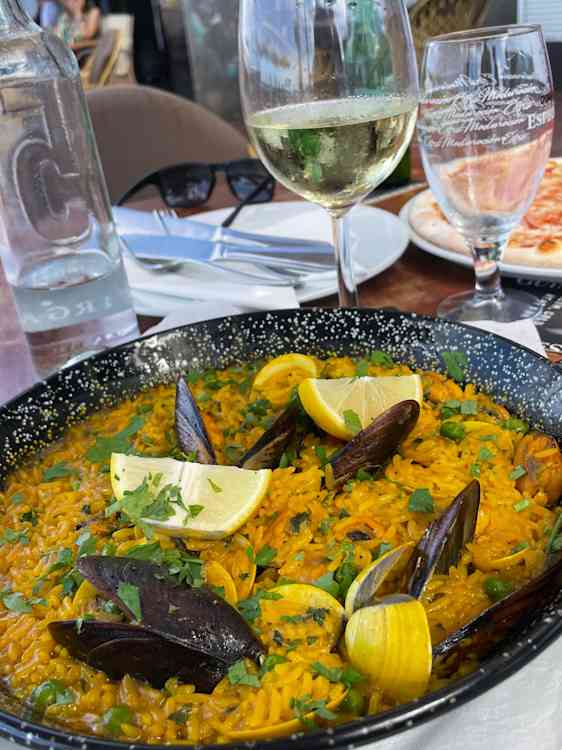 En klassisk paella. En gul risrett med blåskjell, sitronbåter og grønne urter. Dekket bord synes i bakgrunnen.