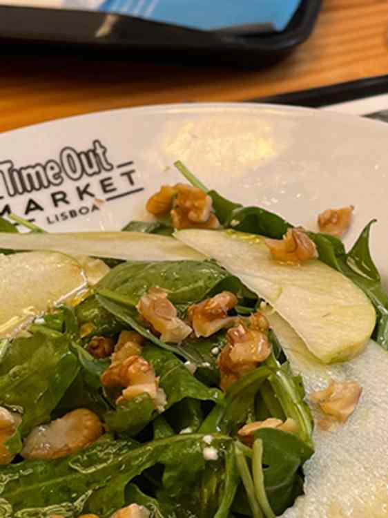 Frisk salat med grønne blader, valnøtter, tynne pæreskiver og revet ost er servert på en hvit tallerken. Tallerkenen står på et trebord og har "Time Out Market Lisboa" trykket på siden.