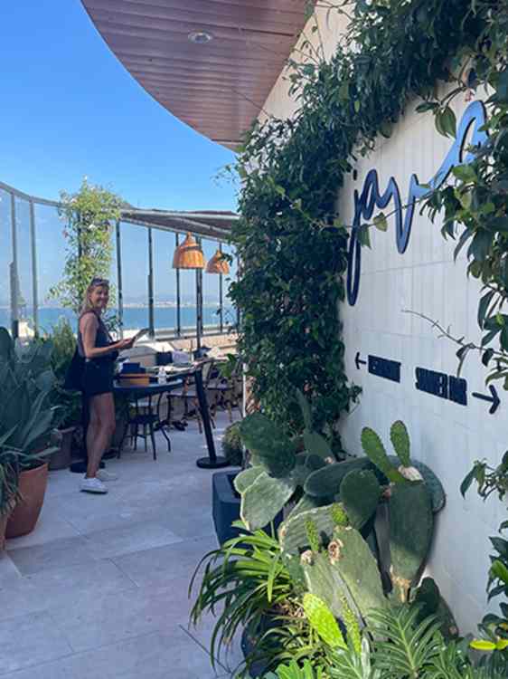 Grønne klatreplanter og kaktuser omgir inngangen til restauranten «Java». Terrassen har bord og stoler med utsikt mot havet under en skyfri himmel.