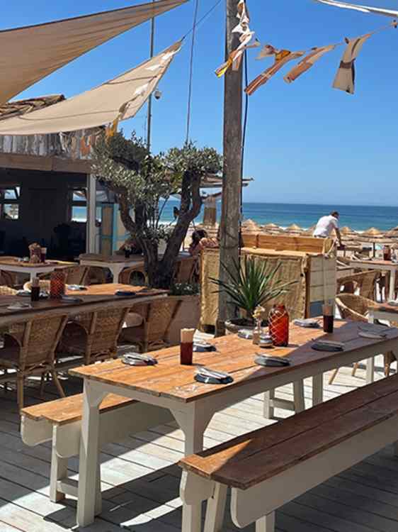 Trebord og benker står på en terrasse med utsikt mot stranden. Seilduk gir skygge, mens havet og stråparasollene er synlige like utenfor serveringsområdet.