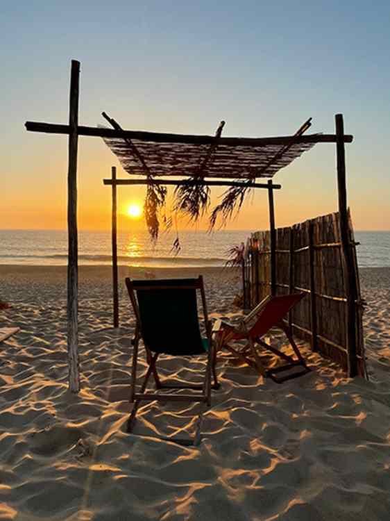 En enkel pergola med flettet tak og strandstoler står på sanden vendt mot havet. Solnedgangen skinner gjennom konstruksjonen og gir stranden et gyllent kveldslys.