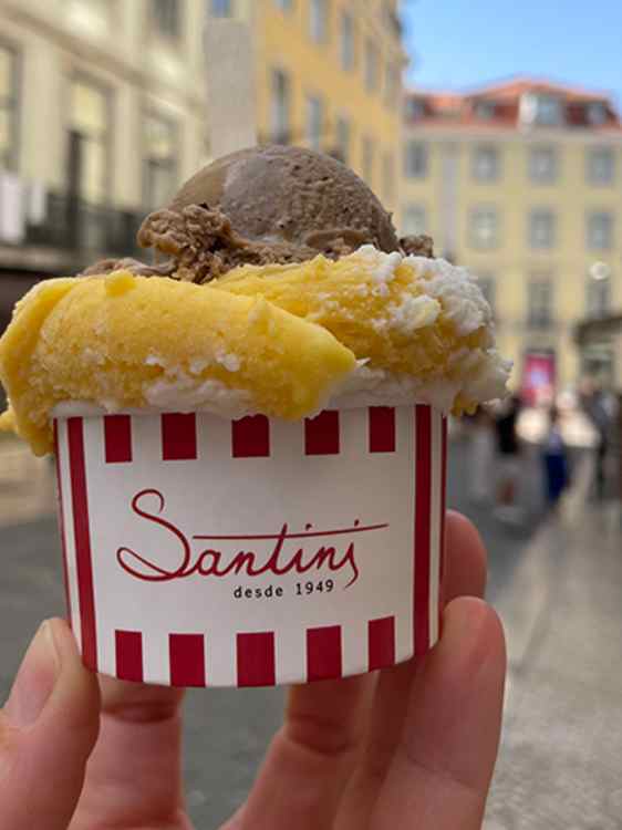Et rødhvitt beger med Santinis logo er fylt med iskrem i flere smaker. Begeret holdes opp foran en solbelyst bygate med uskarpe bygninger i bakgrunnen.