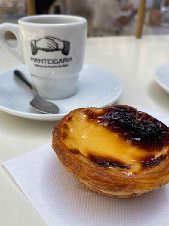 Bakverk med gyllen butterdeig og karamellisert vaniljekrem ligger på en serviett ved siden av en kaffekopp med Manteigarias logo.