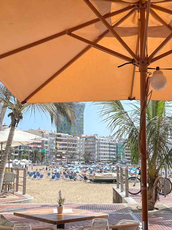 Utsikt fra en terrasse under en stor oransje parasoll mot en bred sandstrand. Blå solsenger og parasoller står spredt utover sanden, med palmer og lyse bygninger langs stranden.