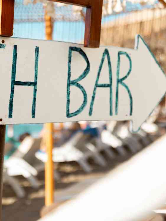 Et hvitt, pilformet skilt med teksten «H BAR» henger foran et uteområde med stoler og bord i bakgrunnen.