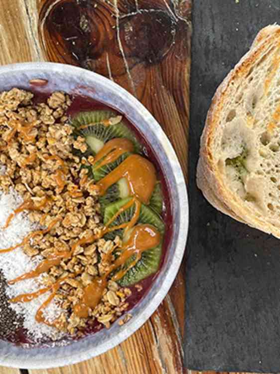 En frokostbolle med granola, skivet kiwi, kokosmasse, chiafrø og en flytende topping står på et rustikt trebord. En stor brødskive ligger ved siden av, sammen med et skjærebrett i bakgrunnen.