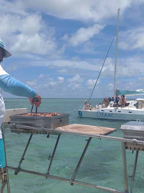 Grillmat tilberedes ved vannkanten, med en seilbåt og personer ute på det turkise havet i bakgrunnen.