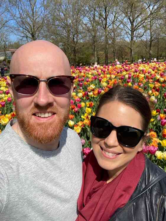 To smilende personer med solbriller står tett sammen foran et stort blomsterbed fylt med røde, gule, rosa og lilla tulipaner.