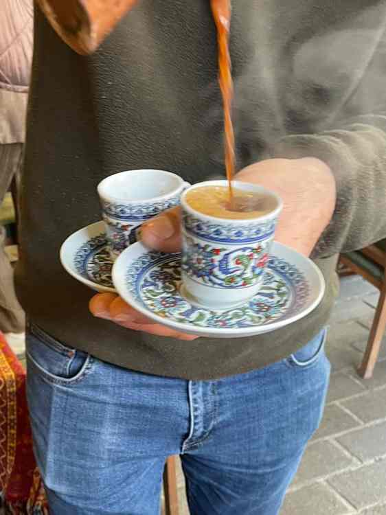 En person holder en kopp med fargerikt blomstermønster på en matchende skål, mens kaffe helles fra en kobberkanne.