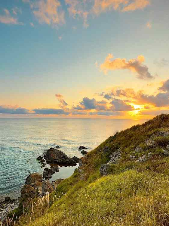 Solen står lavt over havet og lyser opp skyene i gule og oransje nyanser. I forgrunnen skråner en grønn gresskledd åsside ned mot store steiner langs vannkanten.
