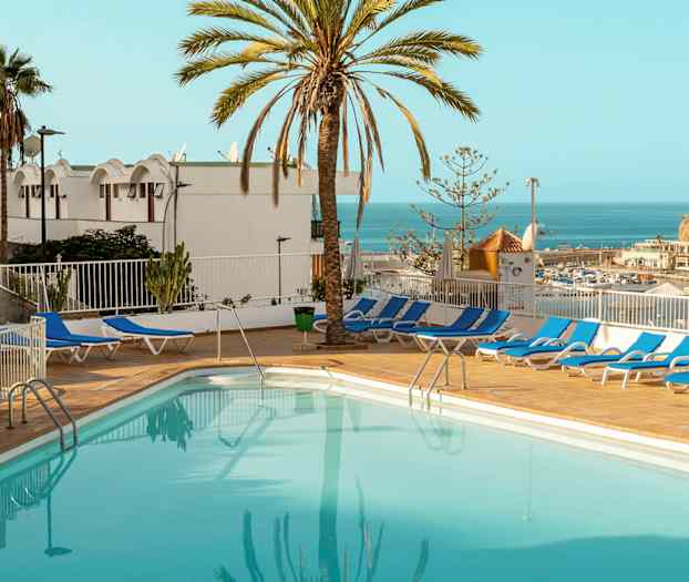 Et utendørs svømmebasseng omgitt av solsenger, med utsikt over havet og omgitt av palmer. (AI-generert tekst)