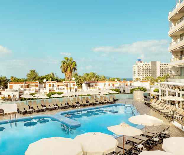 Utsikt over hotellbasseng med solsenger og parasoller, omgitt av bygninger og palmetrær. (AI-generert tekst)