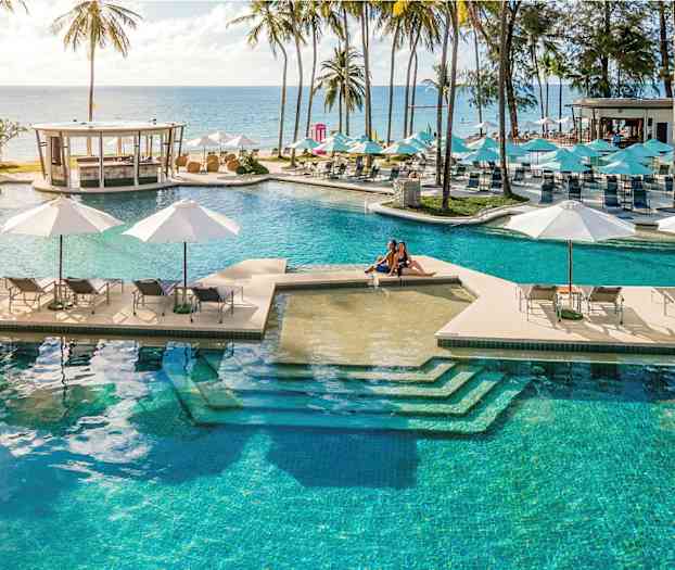 Et stort basseng med solsenger og parasoller med utsikt mot havet, omgitt av palmer. (AI-generert tekst)