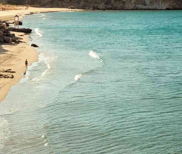 Strand med klart vann og en person som går langs vannkanten. (AI-generert tekst)