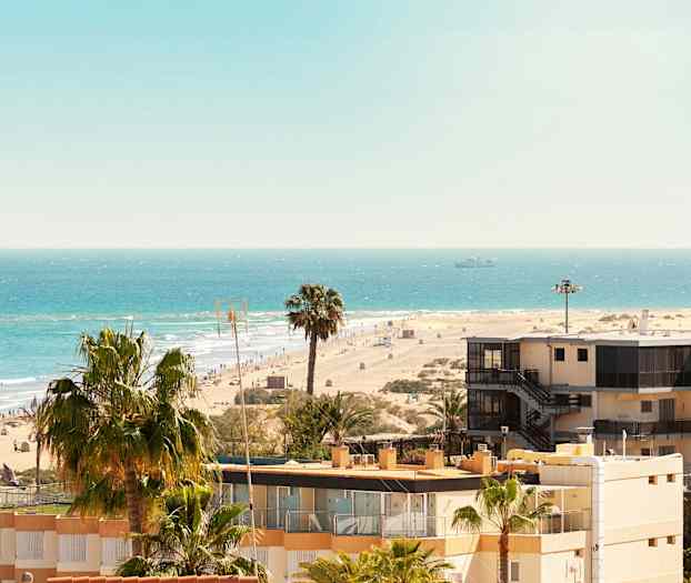 Utsikt over en sandstrand med hav, palmer og bygninger. (AI-generert tekst)