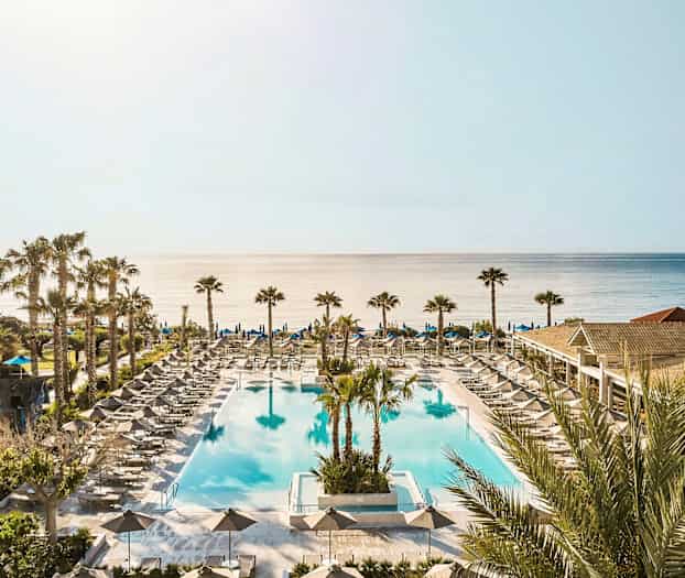 Et stort utendørsbasseng omgitt av solsenger og parasoller med havet i bakgrunnen. (AI-generert tekst)