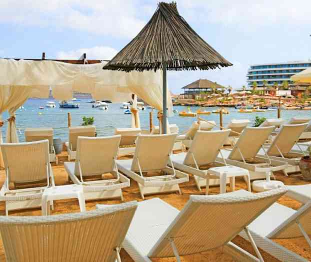 En strand med solsenger foran en rolig havbukt med båter. (AI-generert tekst)