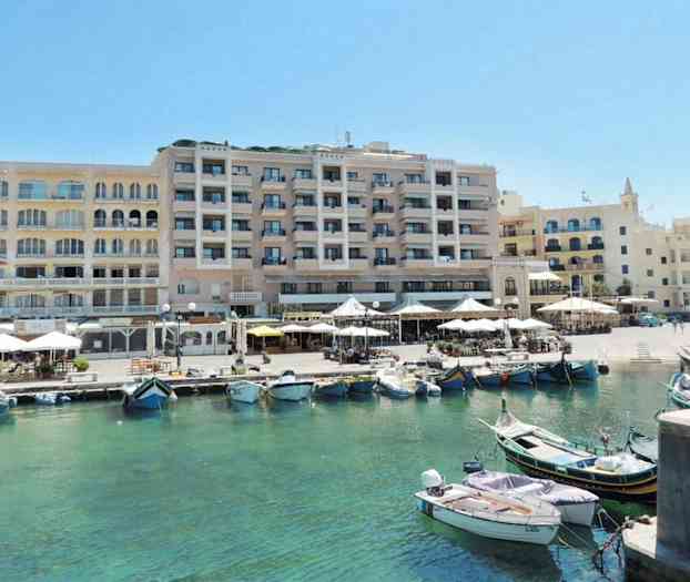 Et bilde av en havn med båter i klart vann foran en rekke bygninger med uteserveringer og terrasser. (AI-generert tekst)