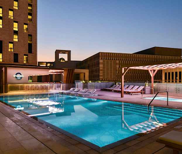 Et opplyst utendørs badebasseng om kvelden omgitt av solsenger og moderne bygningsarkitektur. (AI-generert tekst)