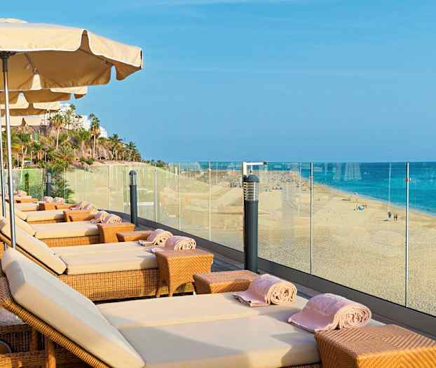 Solstoler med parasoller på en terrasse med utsikt over en sandstrand og havet. (AI-generert tekst)