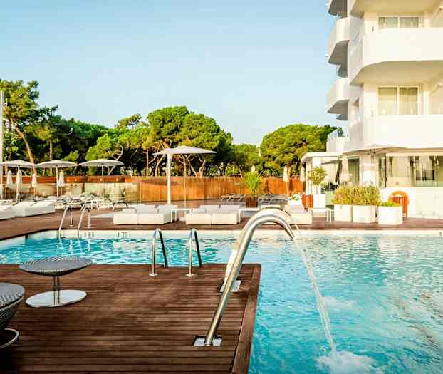 Bilde av et utendørsbasseng ved et hotell, med solsenger og parasoller rundt bassengområdet samt trær i bakgrunnen. (AI-generert tekst)