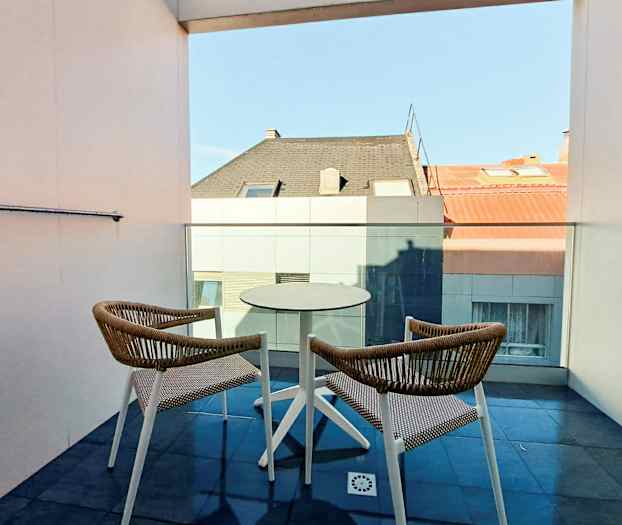 To stoler og et bord på en balkong med utsikt mot hustak. (AI-generert tekst)