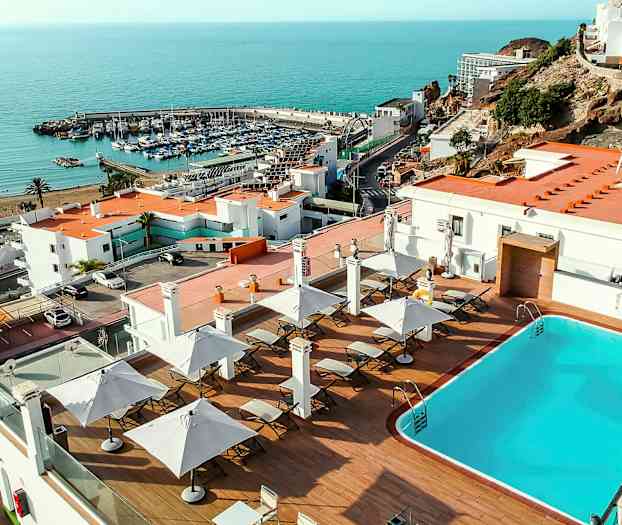 Et basseng med solsenger og parasoller på en terrasse, med utsikt over havet og en marina. (AI-generert tekst)