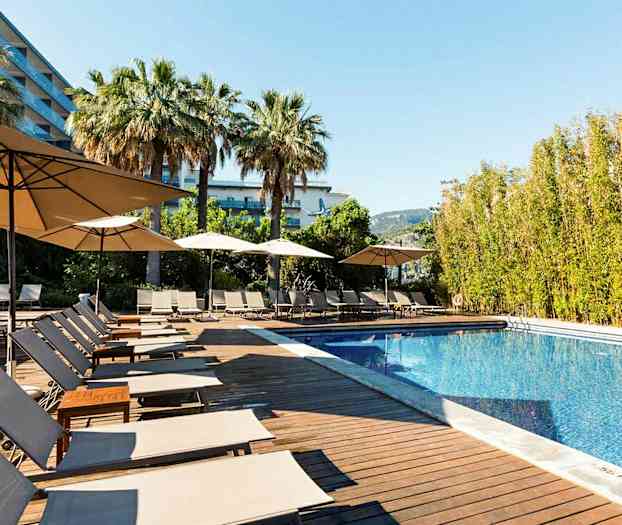 Et utendørs svømmebasseng med solsenger og parasoller omgitt av palmer og trær. (AI-generert tekst)