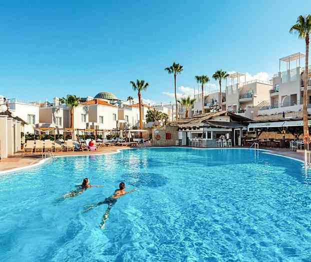 Et stort utendørs basseng foran hvite bygninger og palmer. (AI-generert tekst)