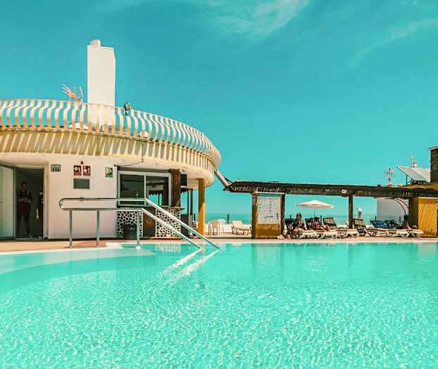 Et utendørs svømmebasseng ved en bygning med solsenger og en klar blå himmel. (AI-generert tekst)