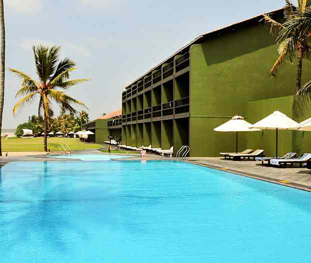 Et utendørs basseng ved et hotell med grønne bygninger, omgitt av palmer og solsenger under parasoller. (AI-generert tekst)