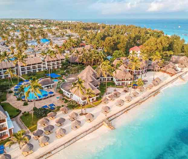 Luftfoto som viser et strandresort med tropiske bygninger omgitt av palmer nær turkist vann. (AI-generert tekst)