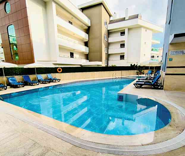 Utendørs basseng med blått vann omgitt av solsenger og bygninger. (AI-generert tekst)