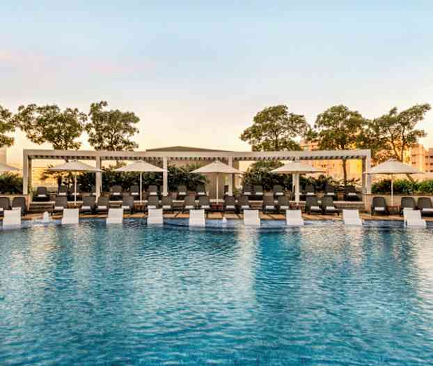 Utendørs svømmebasseng omgitt av solsenger og parasoller med trær i bakgrunnen. (AI-generert tekst)