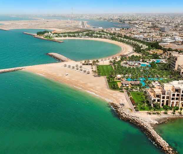 Et flyfoto av en kystby med en halvsirkelformet strand, bygninger, palmer og turkis havvann. (AI-generert tekst)